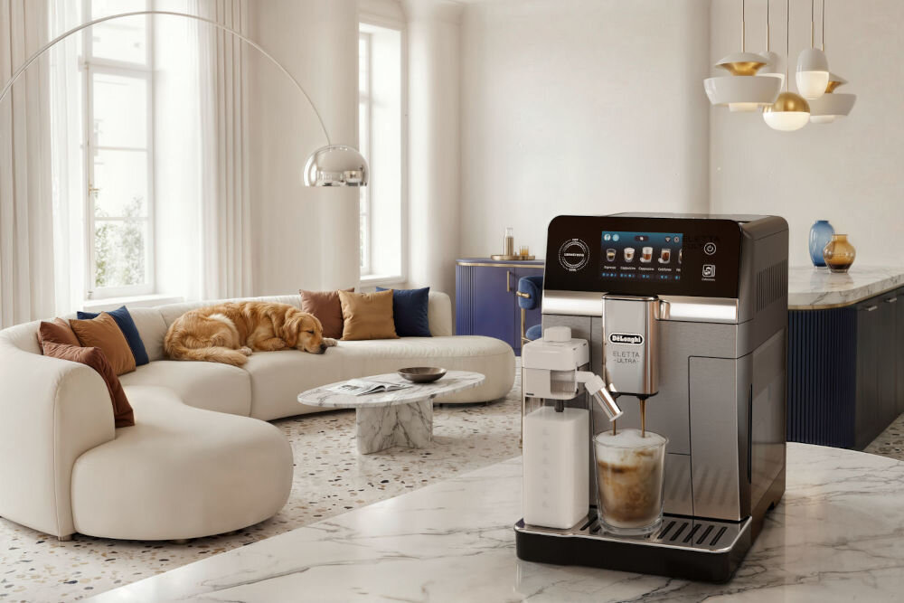 W eleganckim wnętrzu znajduje się ekspres do kawy DELONGHI ECAM470.85.MB Eletta Ultra z funkcją spieniania mleka. Na blacie obok ekspresu stoi szklanka z kawą, a w tle widać sofę z psem oraz stolik kawowy.