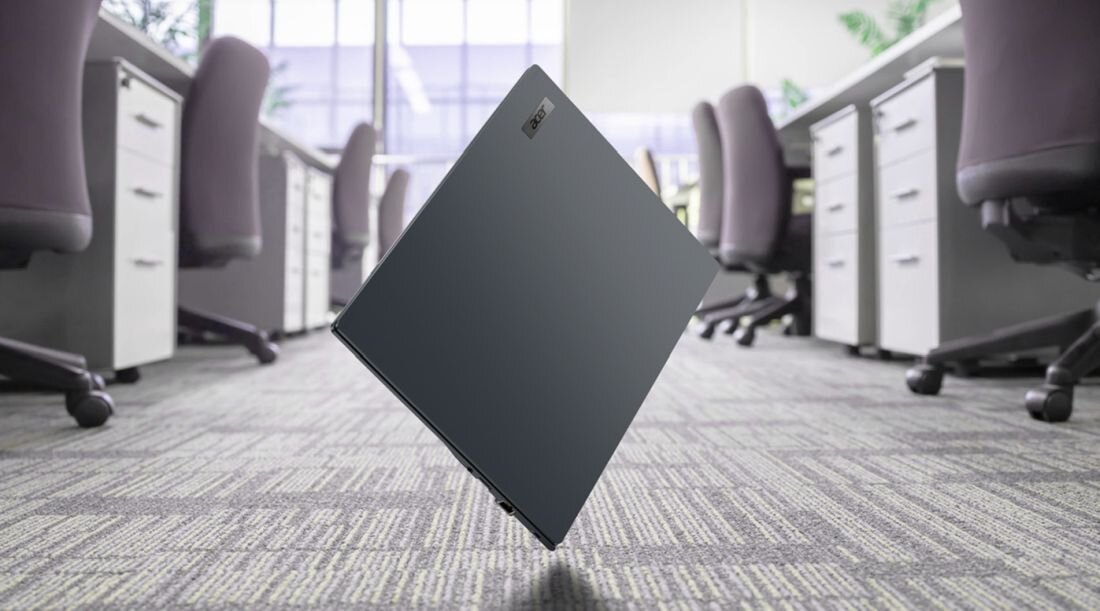 Laptop w kolorze ciemnoszarym unosi się nad podłogą w biurze z rzędem biurek i krzeseł. Na obudowie widoczne jest logo \'ACER\'.
