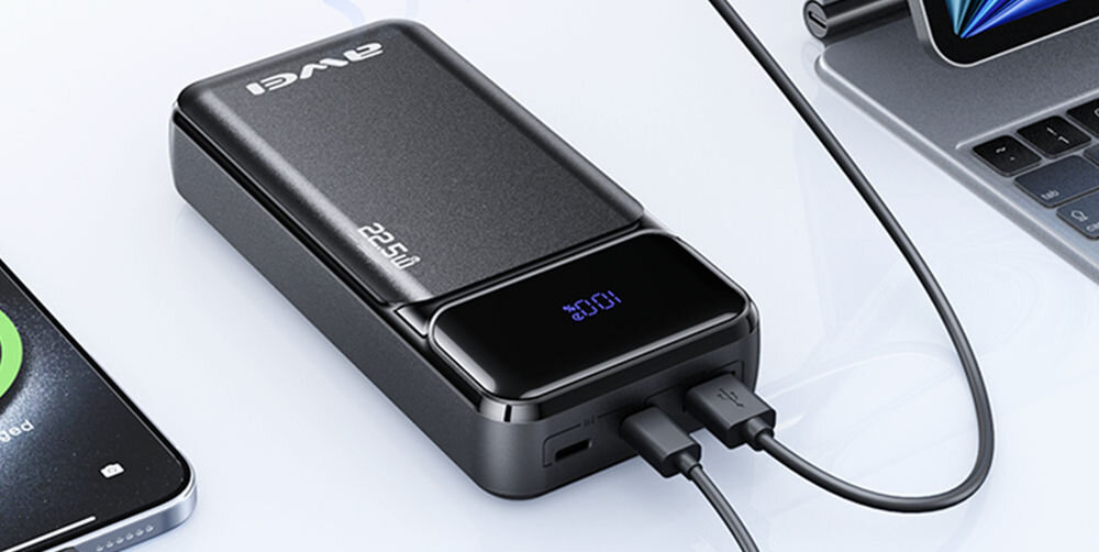Czarny powerbank z logo \'AWEI\' i oznaczeniem \'22.5W\' na obudowie, z wyświetlaczem pokazującym \'100%\'. Urządzenie jest podłączone do dwóch kabli USB, a w tle widać fragmenty smartfona i laptopa.