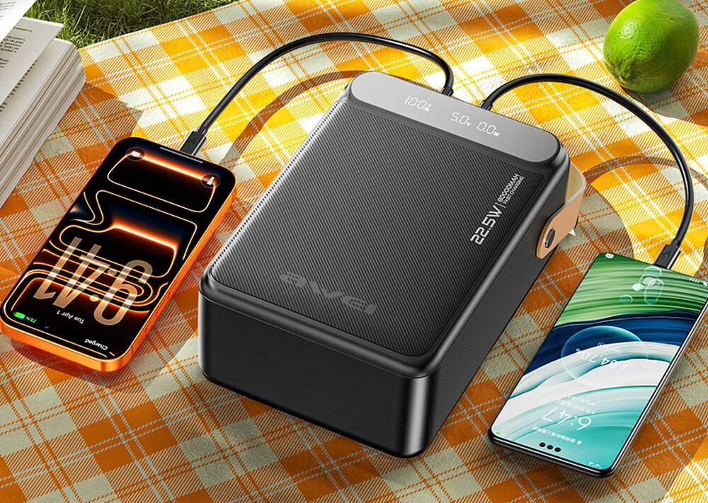 Na obrazie widoczny jest czarny powerbank z napisem \'22.5W PD\' oraz dwa podłączone do niego smartfony. Powerbank leży na pomarańczowym kocu w kratkę, a obok znajduje się limonka.