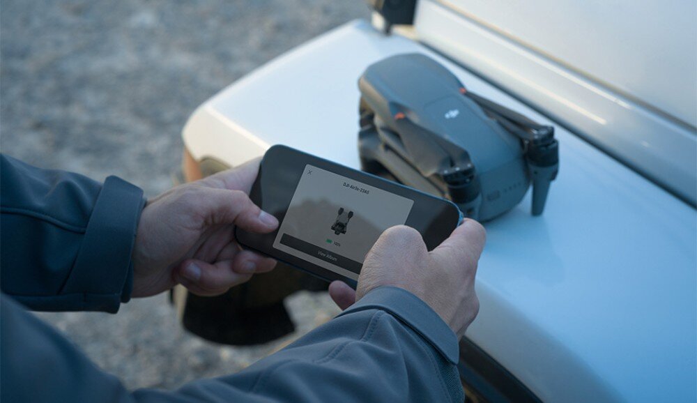 Osoba trzyma kontroler z ekranem, na którym wyświetlany jest obraz drona. Na samochodzie obok leży złożony dron z widocznym logo DJI.