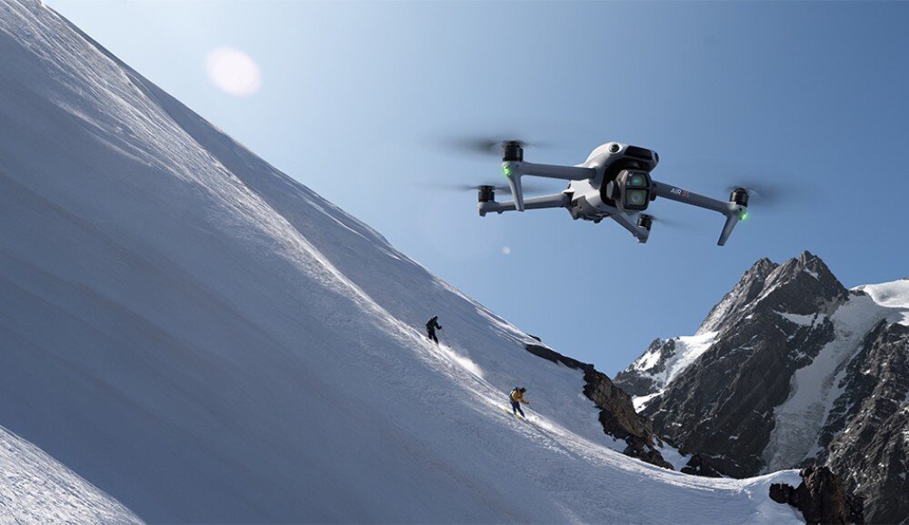 Dron unosi się nad ośnieżonym stokiem górskim, na którym znajdują się dwie osoby. W tle widać szczyty górskie pod błękitnym niebem.