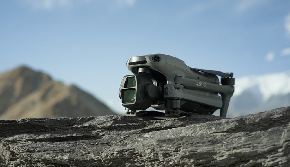 Na obrazie widoczny jest złożony dron w kolorze szarym, umieszczony na kamiennej powierzchni z górami w tle. Na dronie widoczny jest napis \'AIR 3S\'.
