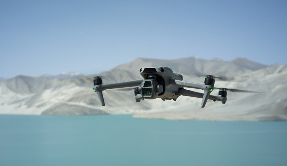 Dron unosi się nad krajobrazem z turkusowym jeziorem i górzystym tłem. Na dronie widoczny jest napis \'AIR\'.