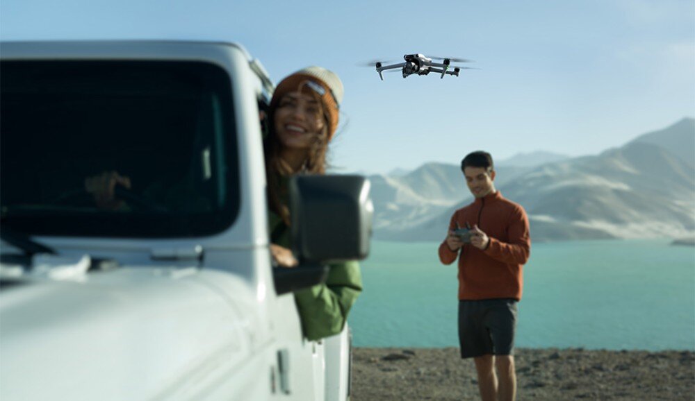 Na pierwszym planie widać samochód terenowy z otwartym oknem, przez które wychyla się osoba. W tle nad jeziorem i górami unosi się dron, a obok stoi osoba trzymająca kontroler.