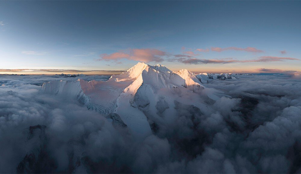 Widok na ośnieżone szczyty górskie otoczone chmurami, oświetlone ciepłym światłem zachodzącego słońca. Na niebie widoczne są delikatne różowe chmury.