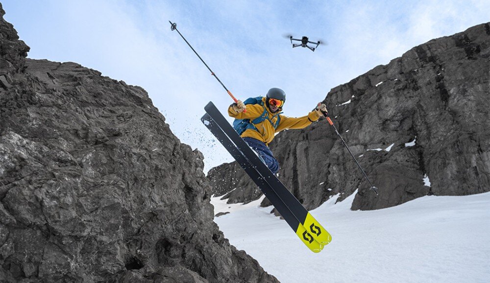 Osoba na nartach skacze z ośnieżonego zbocza górskiego, a nad nią unosi się dron. W tle widać skaliste formacje i śnieg.