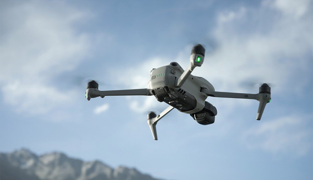 Na obrazie widoczny jest dron w locie na tle górskiego krajobrazu. Dron ma cztery śmigła i zielone światła na ramionach.