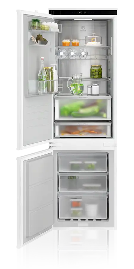 Otwarta lodówka z widocznymi półkami i szufladami, wypełniona różnymi produktami spożywczymi, takimi jak napoje, warzywa i słoiki. Na drzwiach znajdują się dodatkowe półki z butelkami i pojemnikami.