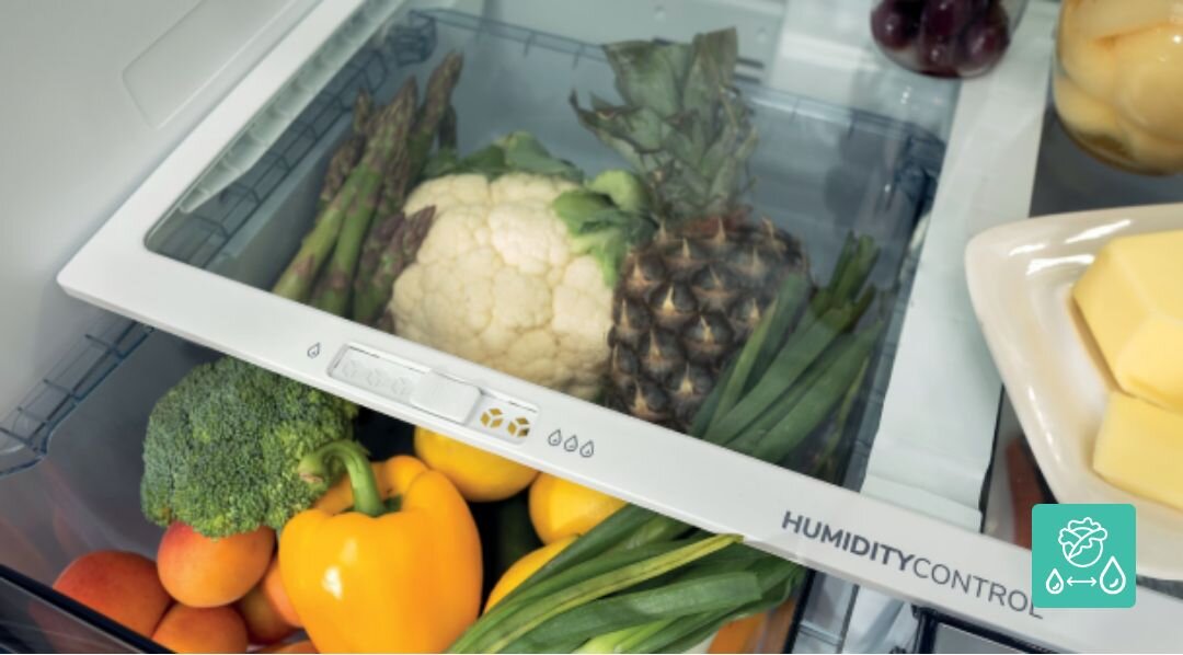 Wnętrze lodówki z szufladą pełną warzyw i owoców, w tym kalafior, ananas, brokuł, papryka i szparagi. Na szufladzie widoczny napis \'HUMIDITY CONTROL\'.