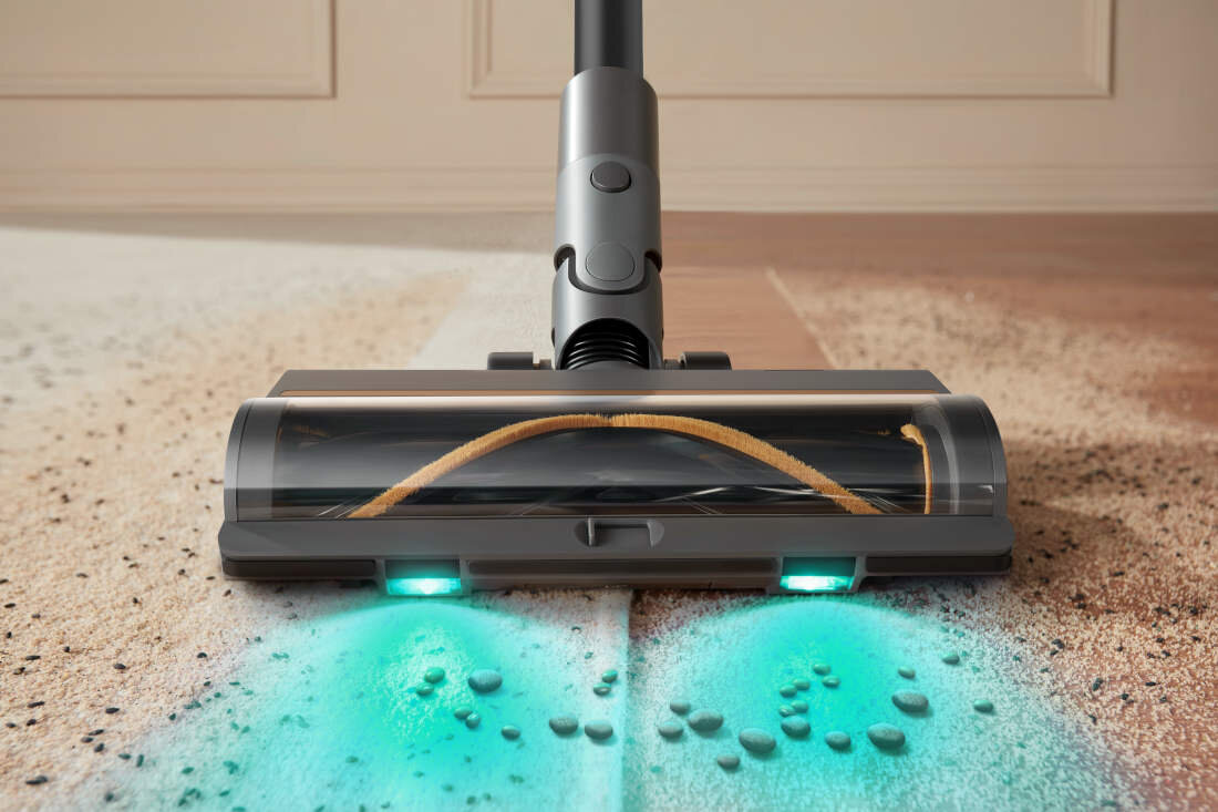 Na obrazie widoczna jest głowica odkurzacza z podświetleniem, która zbiera zanieczyszczenia z podłogi. Podłoga jest częściowo pokryta drobnymi cząstkami, a odkurzacz pozostawia czystą powierzchnię.