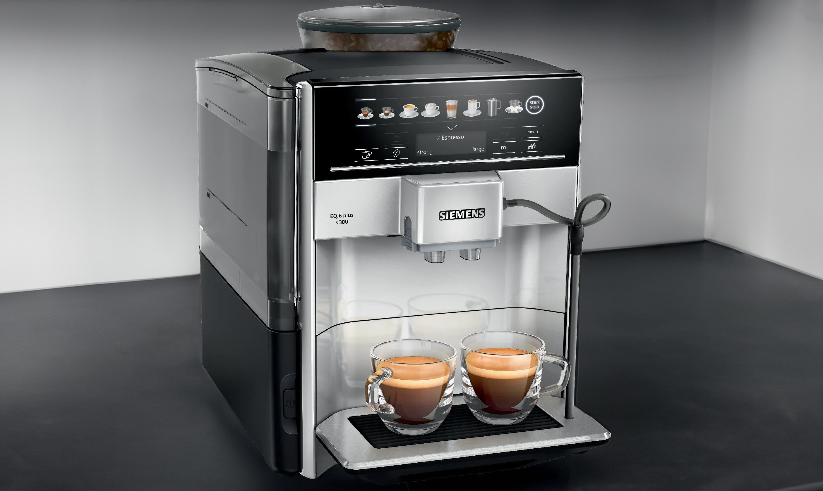 Srebrny ekspres do kawy z wyświetlaczem dotykowym, na którym widoczne są ikony różnych napojów. Na tacy ociekowej stoją dwa szklane kubki z kawą, a na froncie urządzenia znajduje się logo \'SIEMENS\'.