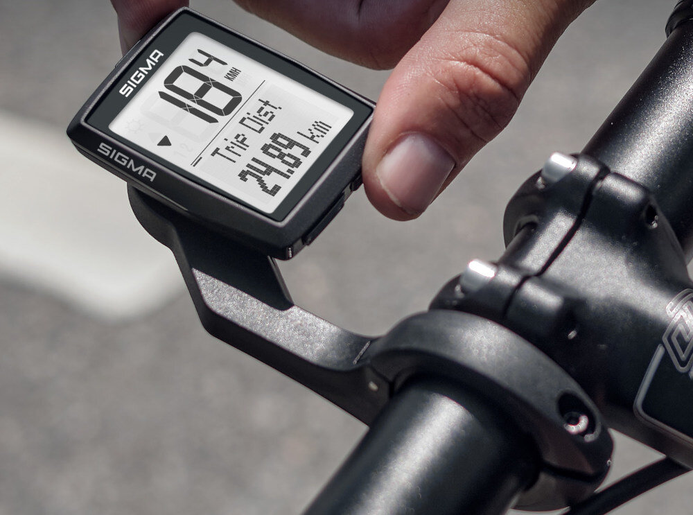 Licznik rowerowy SIGMA zamontowany na kierownicy roweru, wyświetlający prędkość 18.4 km/h oraz dystans 21.89 km. Obok licznika widoczna jest ręka osoby obsługującej urządzenie.
