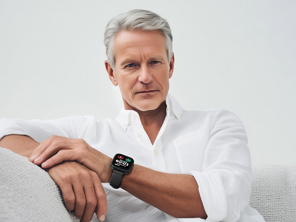 Mężczyzna w białej koszuli siedzi na jasnej kanapie, na jego nadgarstku znajduje się czarny smartwatch z kolorowym wyświetlaczem. Na ekranie zegarka widoczne są ikony i cyfry, ale nie można ich dokładnie odczytać.