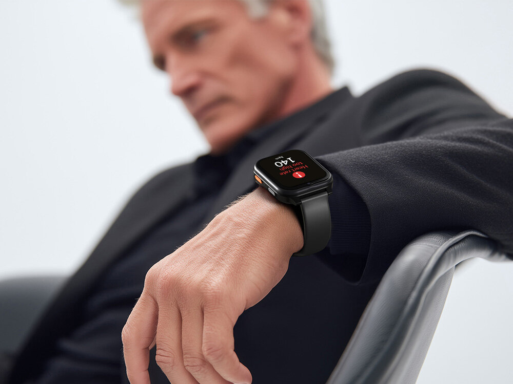 Osoba siedzi na krześle, nosząc czarny smartwatch z wyświetlaczem pokazującym godzinę 2:40 i ikonę telefonu. Tło jest jasne i jednolite.