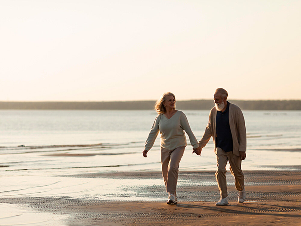 Dwie osoby spacerują po plaży przy zachodzie słońca, trzymając się za ręce. W tle widać spokojne morze i jasne niebo.