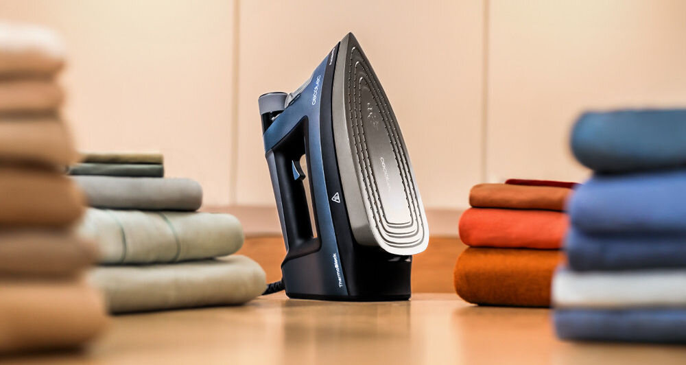 Żelazko w kolorze ciemnym stoi pionowo na drewnianej powierzchni, otoczone stosami kolorowych ubrań. Na żelazku widoczne jest logo \'CECOTEC\'.