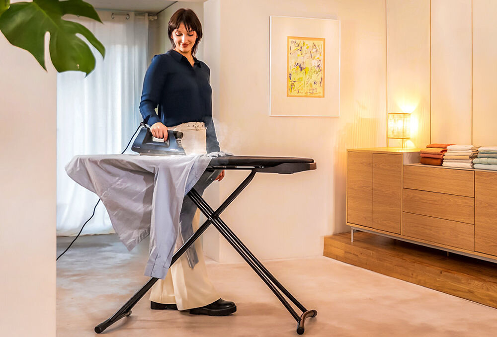 Osoba prasuje koszulę na desce do prasowania w jasnym, nowoczesnym wnętrzu. W tle znajduje się komoda z lampką i ułożonymi ręcznikami.