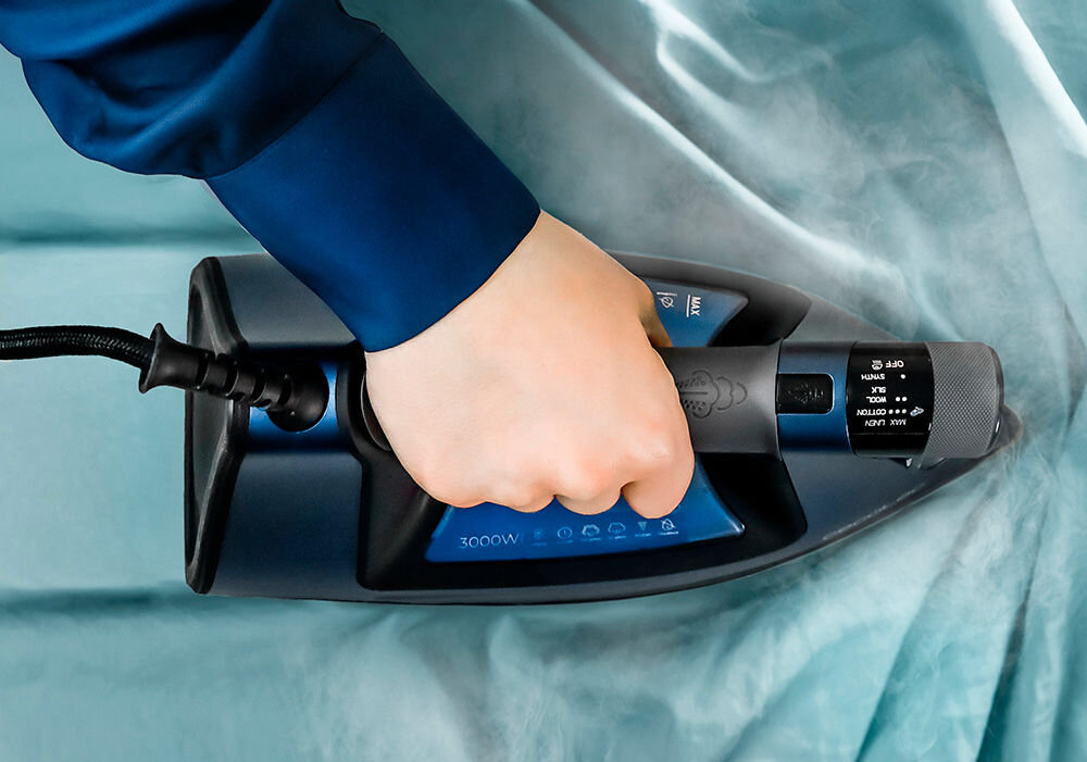Osoba trzyma żelazko na niebieskim materiale, z widocznym napisem \'3000W\' na urządzeniu. Żelazko emituje parę, a na pokrętle widoczne są oznaczenia trybów pracy.