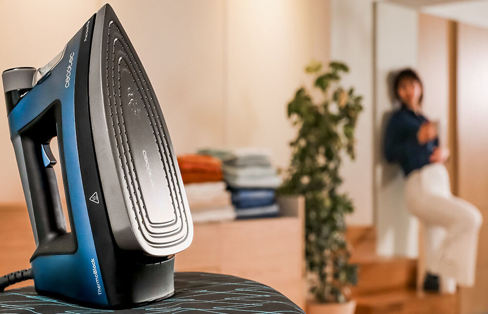 Żelazko CECOTEC z widoczną stopą grzewczą i logo \'cecotec\' na desce do prasowania. W tle znajduje się osoba oraz stos złożonych ręczników.