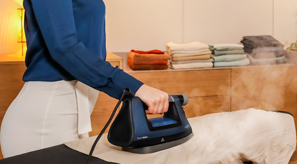 Osoba prasuje tkaninę za pomocą żelazka, z którego wydobywa się para. W tle widoczne są złożone ręczniki na drewnianej komodzie.