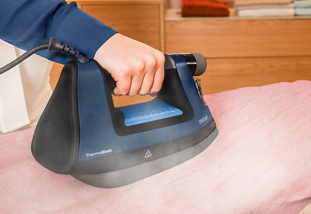 Osoba prasuje różowy materiał za pomocą niebieskiego żelazka z napisem \'ThermoBlock\' i \'CECOTEC\'. Z żelazka wydobywa się para.