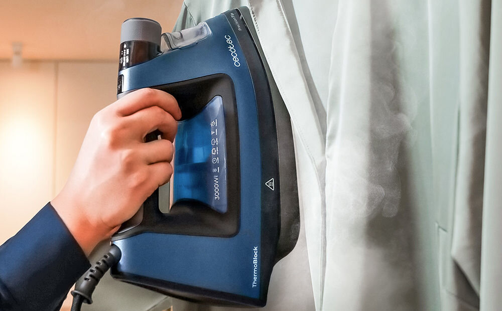 Osoba prasuje ubranie za pomocą niebieskiego żelazka z widocznym napisem \'Cecotec\' oraz oznaczeniami \'3000W\' i \'ThermoBlock\'.