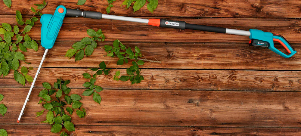 Na drewnianym tle leżą akumulatorowe nożyce do żywopłotu z długim uchwytem, w kolorze niebieskim i czarnym. Widoczne jest logo \'GARDENA\' na uchwycie.