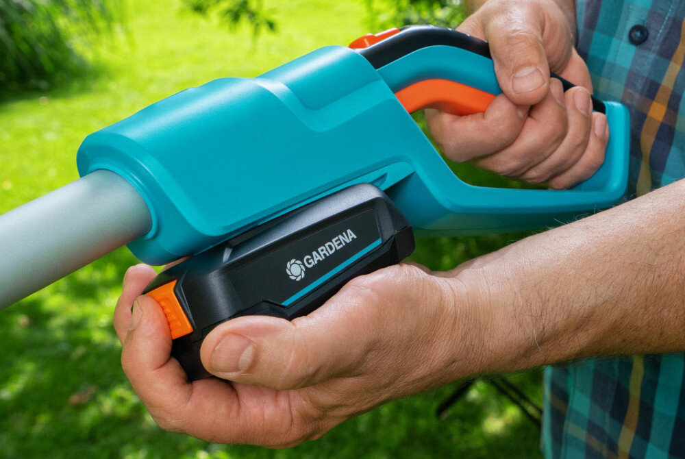 Osoba trzyma niebiesko-pomarańczowe nożyce do żywopłotu z widocznym logo \'GARDENA\'. W tle znajduje się zielona trawa.