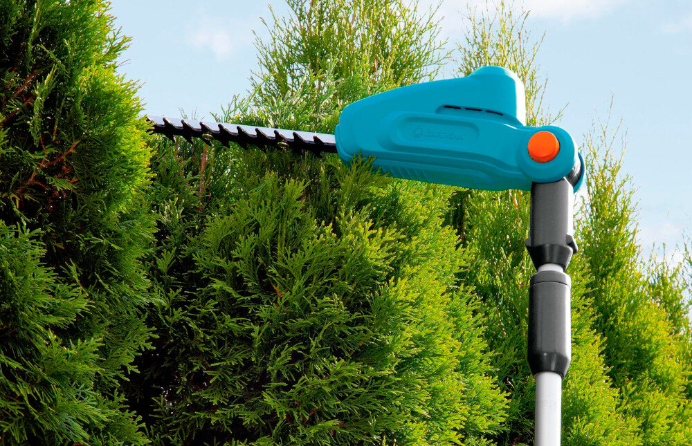 Na obrazie widoczne są nożyce do żywopłotu z niebieską obudową i czarnym uchwytem, używane do przycinania zielonego żywopłotu. Na obudowie znajduje się logo \'Gardena\'.