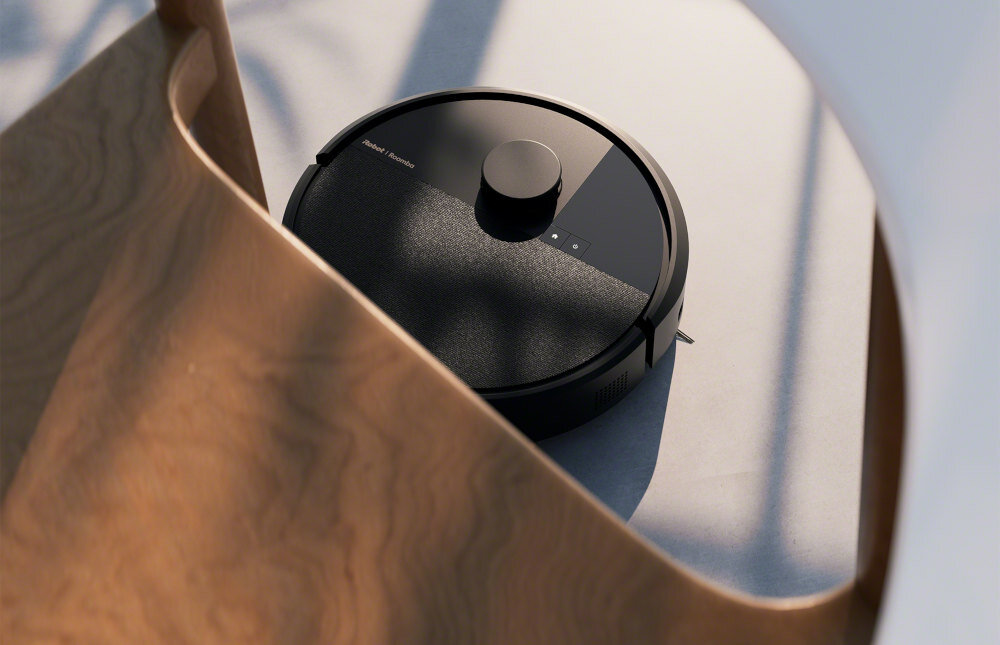 Czarny robot sprzątający z widocznym logo \'IROBOT Roomba\' na górnej części obudowy, umieszczony na jasnej powierzchni w otoczeniu drewnianych elementów.