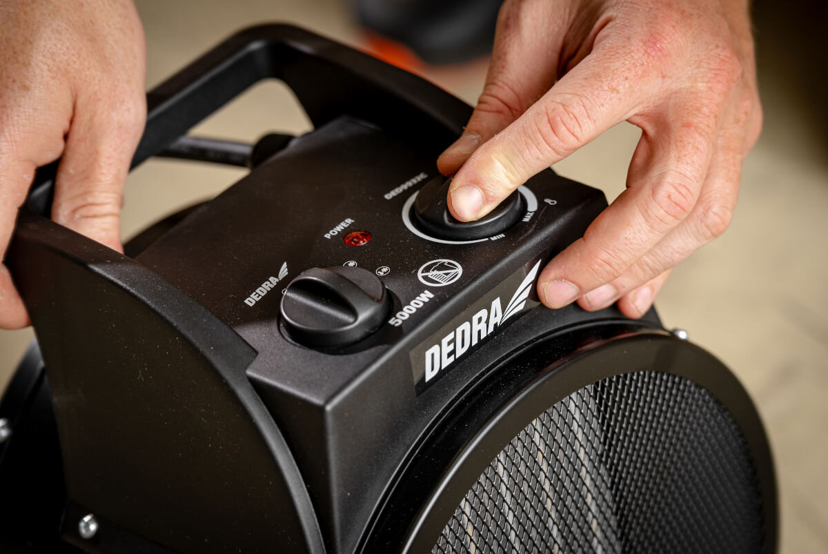 Osoba reguluje pokrętło na czarnej nagrzewnicy elektrycznej z widocznym logo \'DEDRA\'. Na panelu znajdują się napisy: \'POWER\', \'5000W\' oraz symbol wentylatora.
