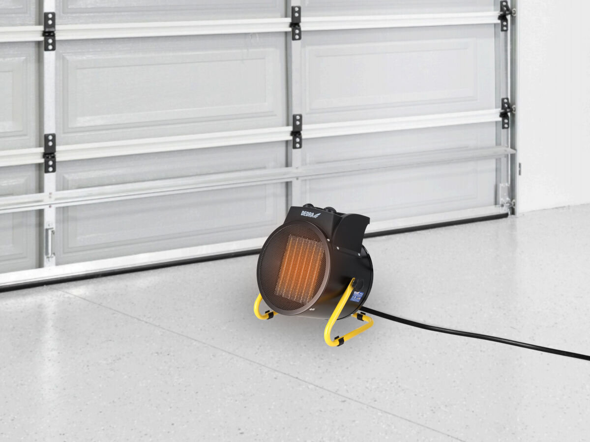 Na podłodze w garażu stoi czarna nagrzewnica elektryczna z żółtym stojakiem i widocznym logo \'DEDRA\'. Urządzenie jest podłączone do prądu za pomocą czarnego kabla.