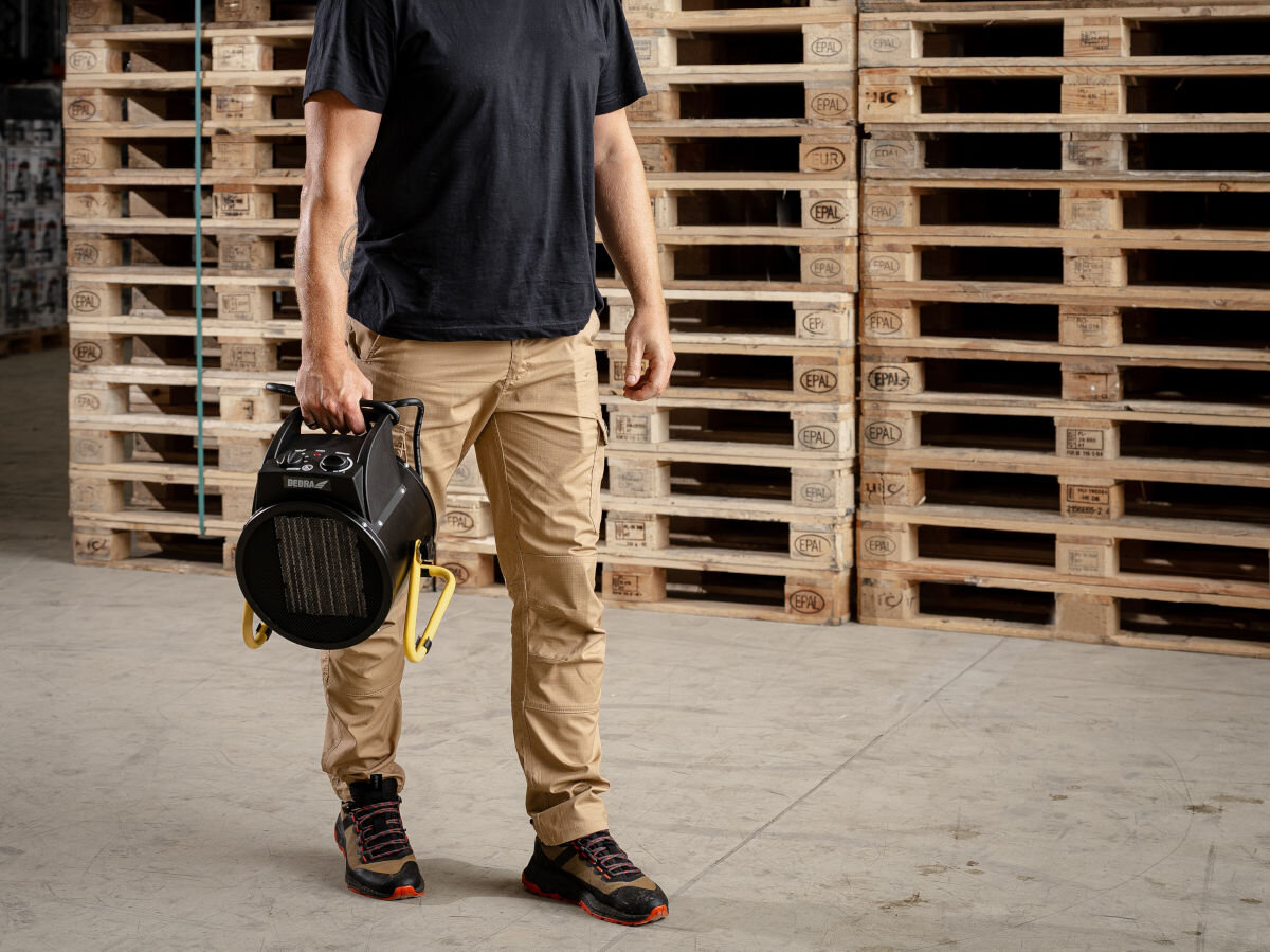 Osoba trzymająca czarną nagrzewnicę elektryczną z żółtymi elementami, stojąca na tle stosu drewnianych palet. Na nagrzewnicy widoczne jest logo producenta.