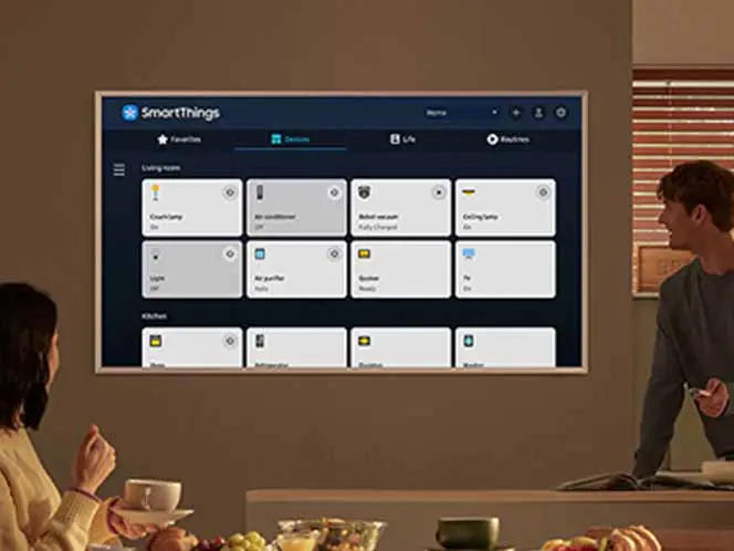 Na ścianie wisi telewizor z wyświetlonym interfejsem aplikacji SmartThings, pokazującym różne urządzenia domowe. W pomieszczeniu znajdują się dwie osoby, jedna siedzi przy stole, a druga stoi obok telewizora.