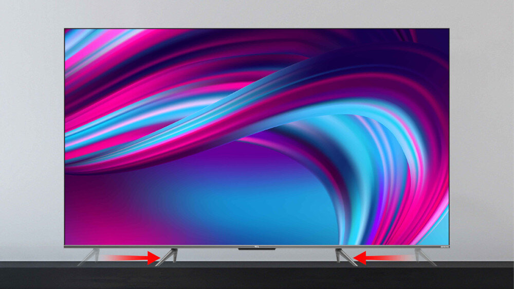 Telewizor z dużym ekranem wyświetlający kolorowe, abstrakcyjne wzory w odcieniach różu i niebieskiego. Na dolnej krawędzi ekranu widoczne są dwie czerwone strzałki skierowane w stronę podstawy telewizora.