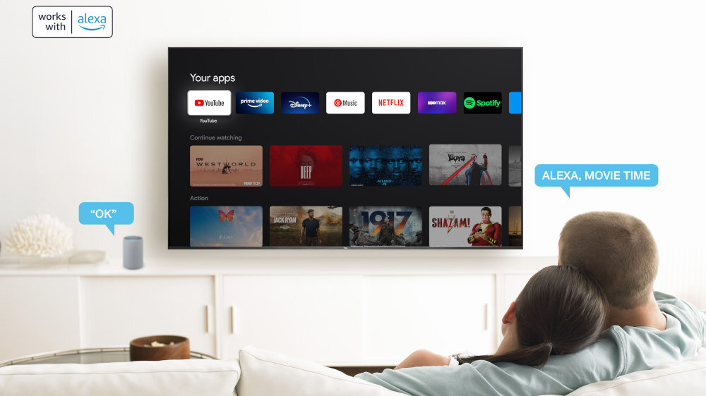 Na obrazie widać telewizor z interfejsem Google TV, na którym wyświetlane są aplikacje takie jak YouTube, Prime Video, Disney+, Music, Netflix i Spotify. Obok telewizora znajduje się napis \'works with alexa\', a na kanapie siedzą dwie osoby.