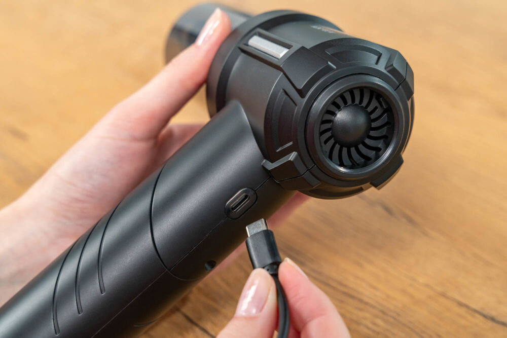 Osoba trzyma czarny odkurzacz ręczny, do którego podłącza kabel z wtyczką USB-C. Odkurzacz ma widoczne detale konstrukcyjne i znajduje się na tle drewnianej powierzchni.