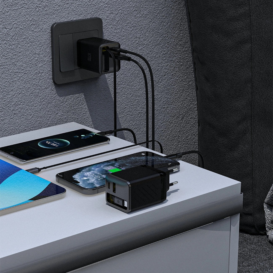 Na zdjęciu widać czarną ładowarkę sieciową podłączoną do gniazdka, z której wychodzą trzy kable USB-C ładujące urządzenia elektroniczne na białym blacie. Obok ładowarki leży dodatkowy adapter z dwoma portami USB.