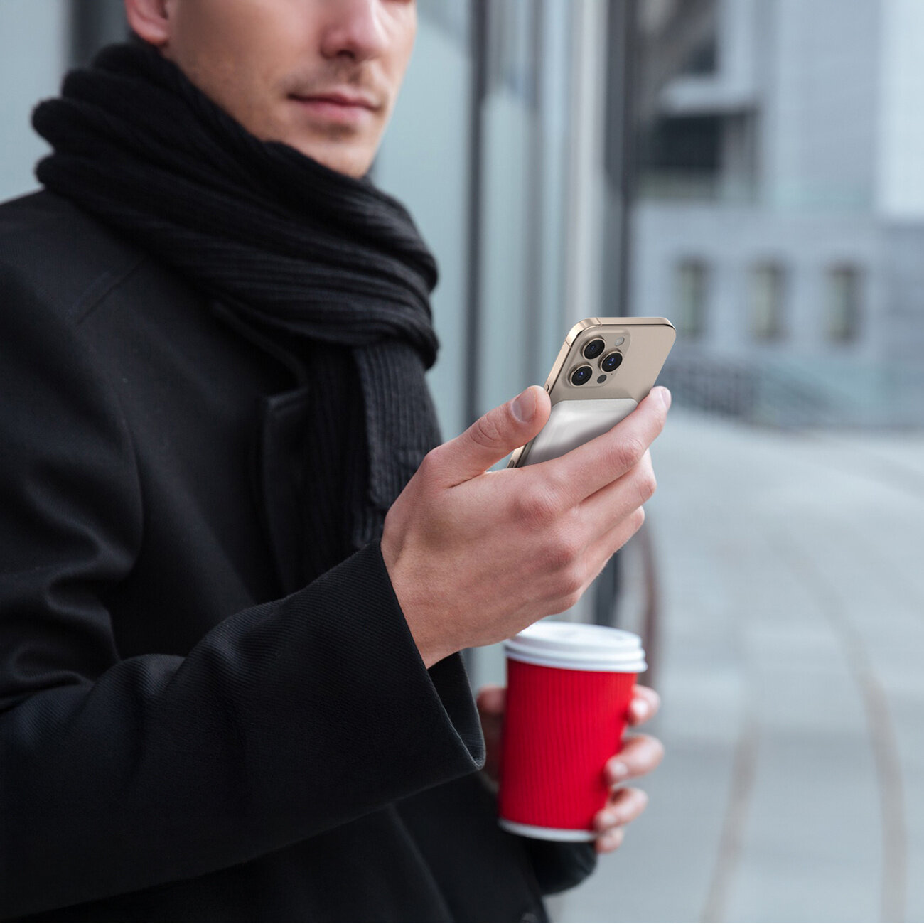 Osoba w ciemnym płaszczu i szaliku trzyma w jednej ręce smartfon, a w drugiej czerwony kubek z napojem. W tle widoczne są szklane ściany budynku.