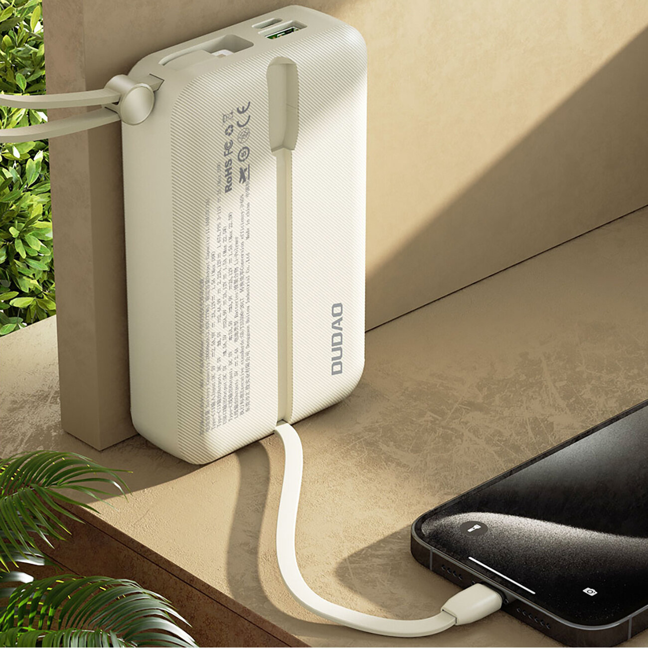 Biały powerbank z logo \'DUDAO\' leży na powierzchni, z podłączonym kablem do smartfona. Na urządzeniu widoczne są porty USB i tekst z informacjami technicznymi.