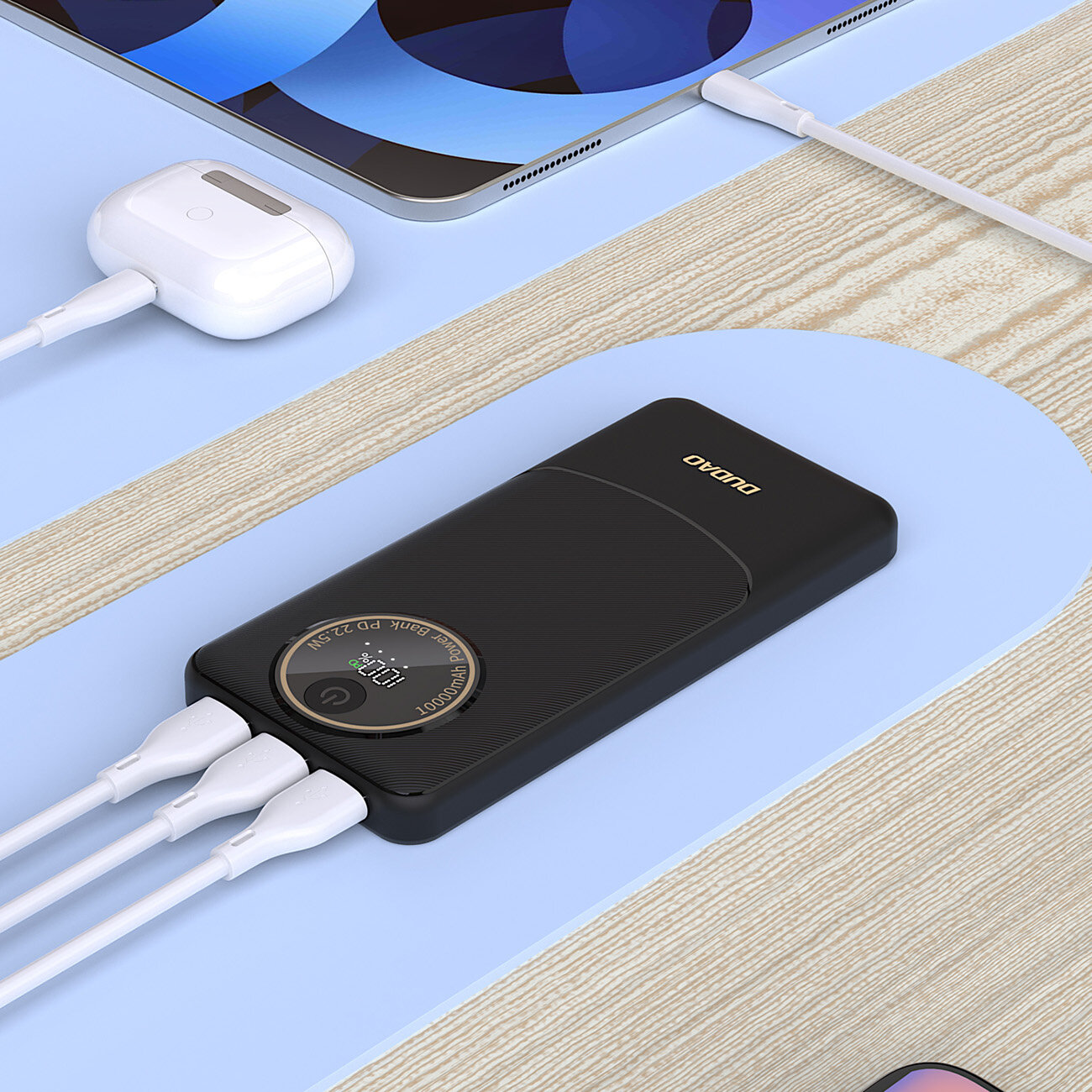 Na obrazie znajduje się czarny powerbank z logo \'DUDAO\', podłączony do trzech białych kabli. Obok widoczne są słuchawki bezprzewodowe i tablet.