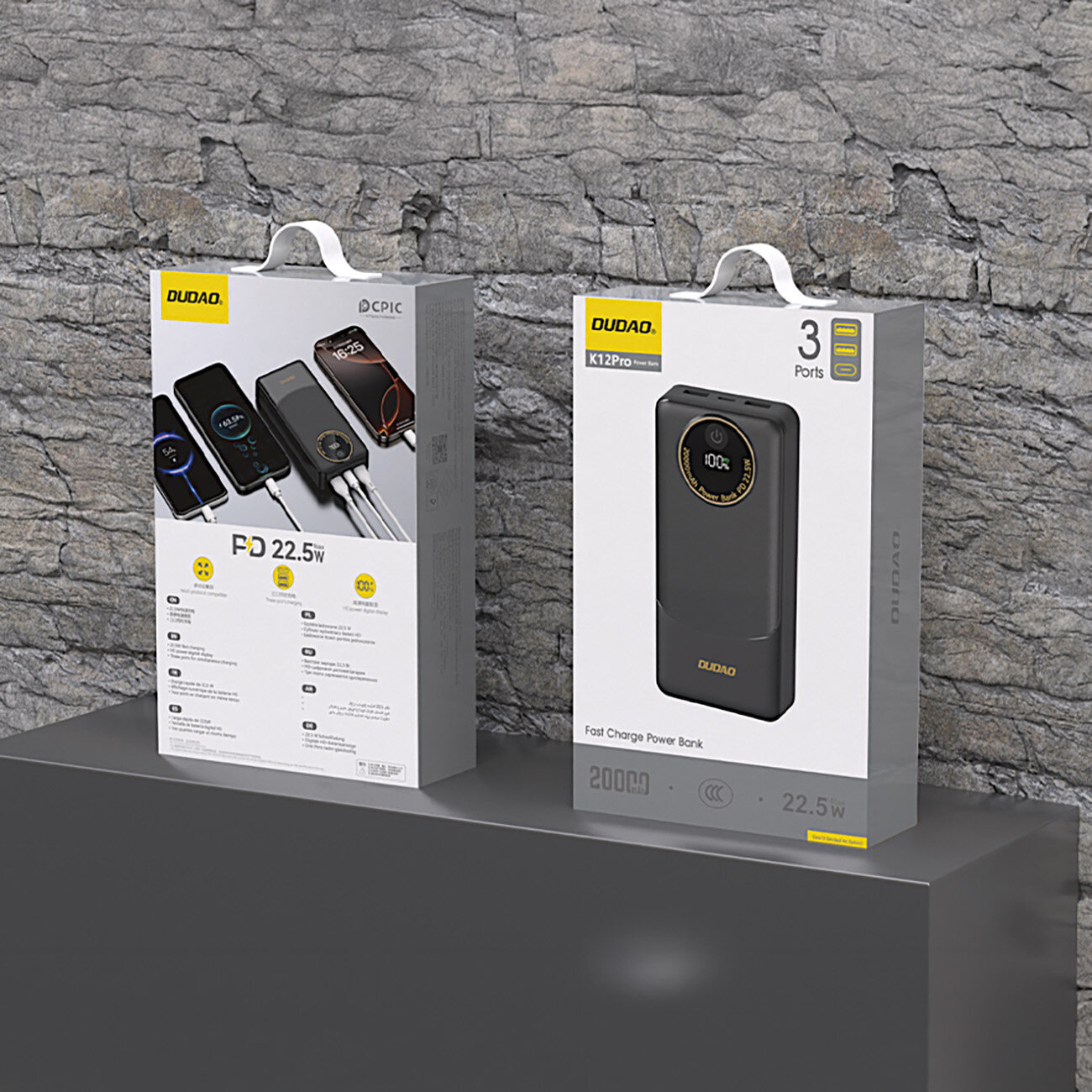 Na obrazie widoczne są dwa opakowania produktów marki Dudao, ustawione na tle kamiennej ściany. Opakowania zawierają informacje o powerbanku, w tym \'K12Pro\', \'3 Ports\', \'20000mAh\', \'22.5W\'.