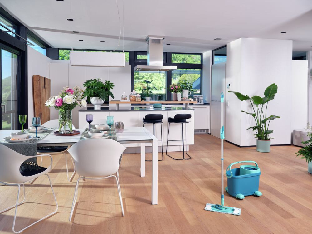 Nowoczesna kuchnia z dużymi oknami, jasnymi meblami i drewnianą podłogą. Na podłodze stoi mop płaski z wiadrem w kolorze turkusowym.