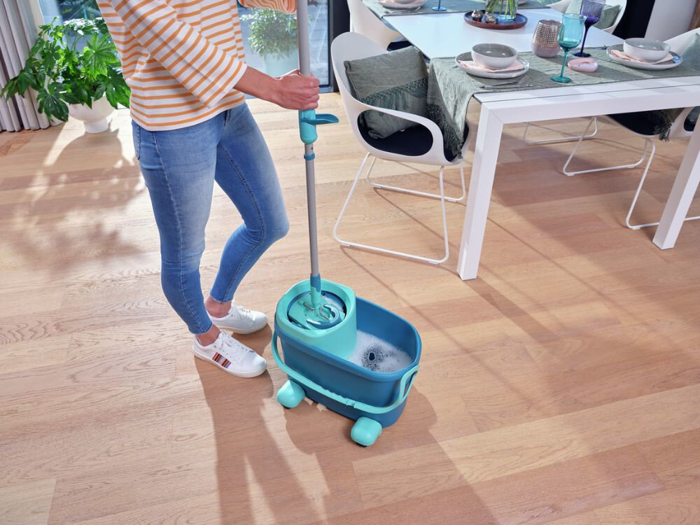 Osoba używa mopa z turkusowym wiadrem na kółkach, wypełnionym wodą z pianą, na drewnianej podłodze w jasnym pomieszczeniu. W tle znajduje się stół z nakryciem i krzesłami.