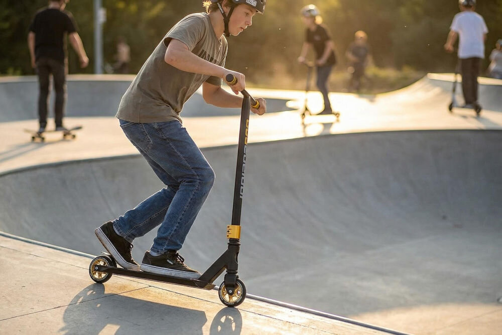 Osoba na hulajnodze wyczynowej w skateparku, w tle widać innych użytkowników na hulajnogach i deskorolkach. Hulajnoga ma czarno-zielony kolor z widocznym napisem \'INDIANA\'.