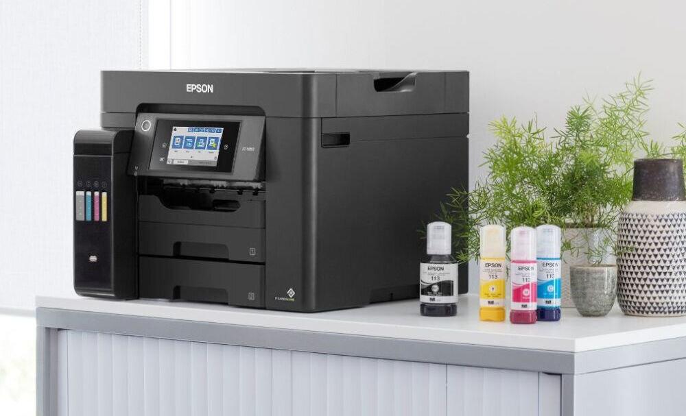 Na obrazie znajduje się czarne urządzenie wielofunkcyjne z logo EPSON, z wyświetlaczem dotykowym i widocznymi pojemnikami na tusz. Obok urządzenia stoją cztery butelki z tuszem w różnych kolorach oraz roślina w doniczce.