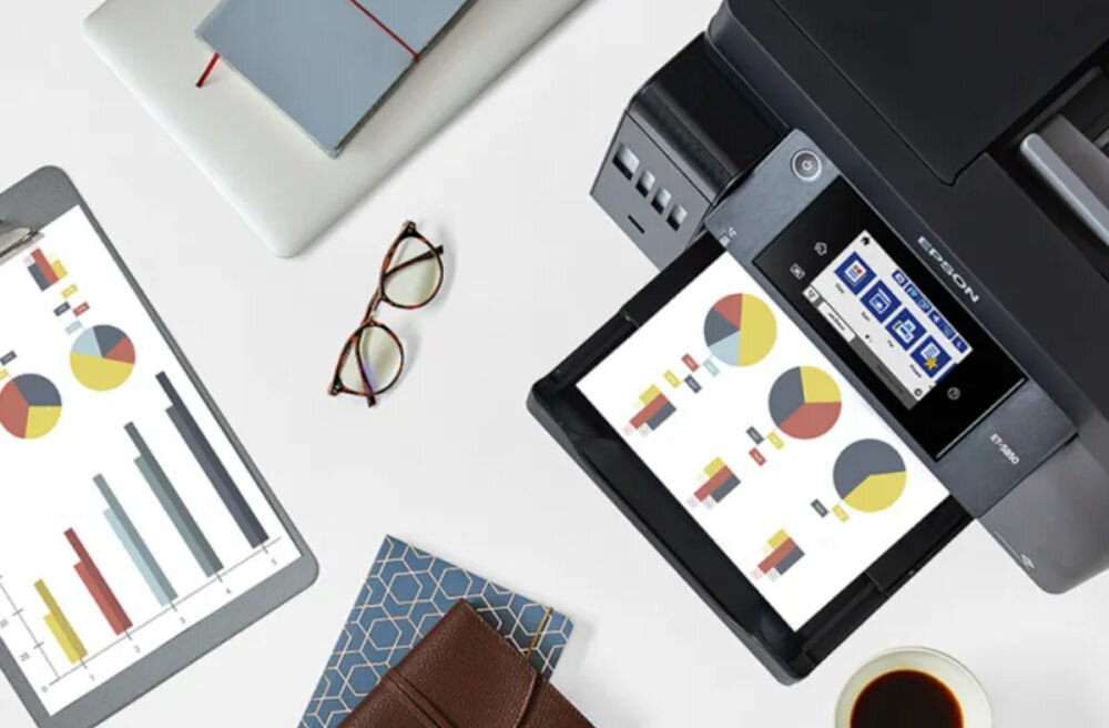 Na biurku znajduje się urządzenie wielofunkcyjne z logo EPSON, które drukuje kolorowy wykres. Obok leżą okulary, tablet z wykresami, notes, filiżanka kawy i laptop.