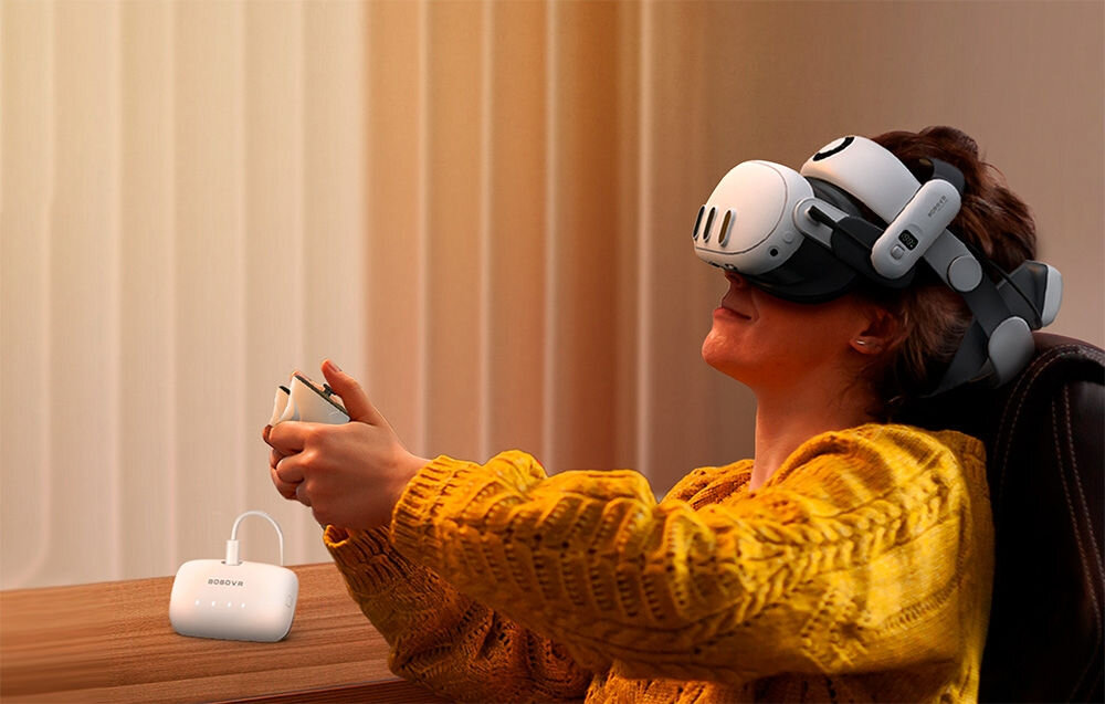 Osoba siedzi na krześle, nosząc zestaw VR, trzymając kontroler w rękach. Obok na stole znajduje się biały akumulator z napisem \'BOBOVR\'.