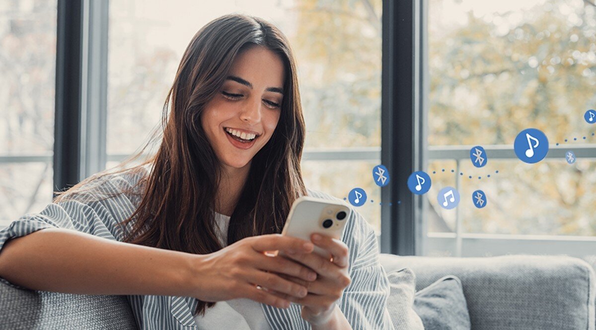 Osoba siedzi na kanapie, trzymając w rękach smartfon. W tle widoczne są ikony Bluetooth i nutki, sugerujące przesyłanie muzyki.